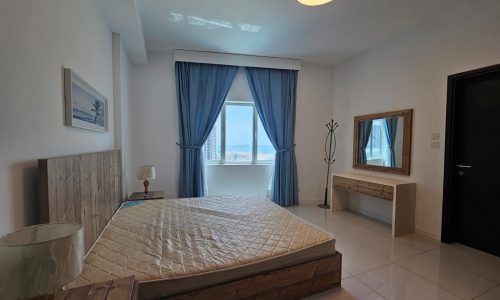 A modern two-bedroom apartment featuring a large bed with a beige comforter, wooden bed frame, a dresser with a mirror, and a window with ocean view.