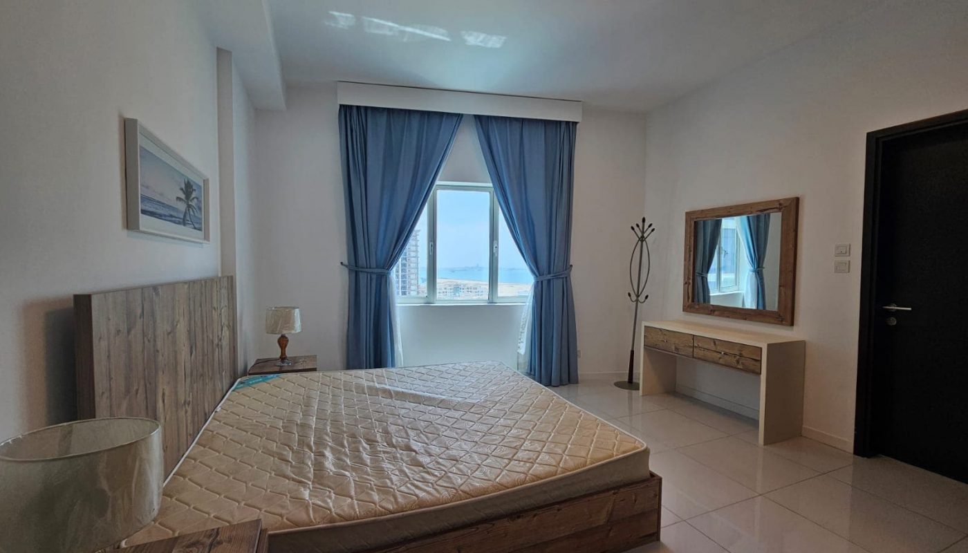 A modern two-bedroom apartment featuring a large bed with a beige comforter, wooden bed frame, a dresser with a mirror, and a window with ocean view.