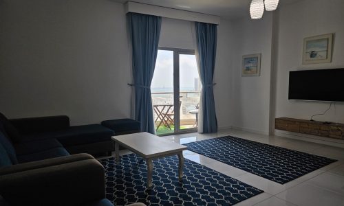 Interior of a fully furnished modern living room in a two-bedroom apartment with a view of the ocean through a large window, featuring a sofa, coffee table, and sea-themed decor.