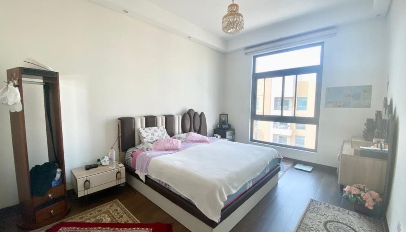 A bedroom with a large bed, wooden floor, area rugs, a window, and a chandelier. There's a side table, a wardrobe, and a dresser.