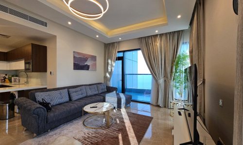 Modern living room with a gray sectional sofa, a circular coffee table, and large windows draped in sheer curtains, leading to a balcony.