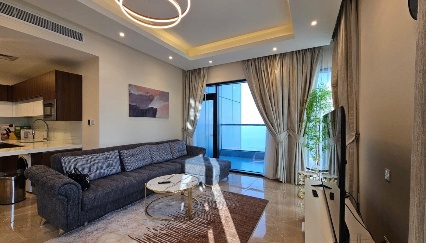 Modern living room with a gray sectional sofa, a circular coffee table, and large windows draped in sheer curtains, leading to a balcony.