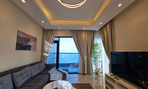 Modern living room with a large sofa, round coffee table, and a view of the ocean through a sliding glass door. bright interior with artistic ceiling lights and a wall painting.