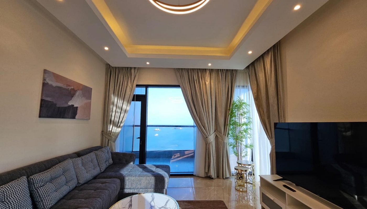 Modern living room with a large sofa, round coffee table, and a view of the ocean through a sliding glass door. bright interior with artistic ceiling lights and a wall painting.