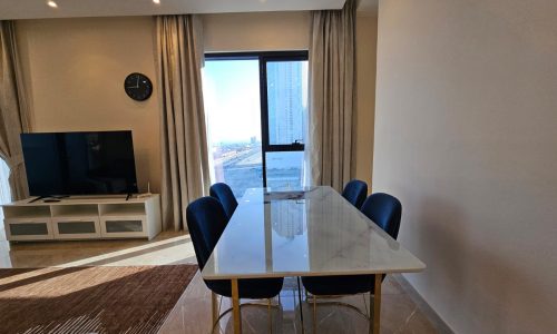 Interior of a modern living room featuring a marble dining table with blue chairs, a tv on a white stand, and a view of the city through a balcony door.