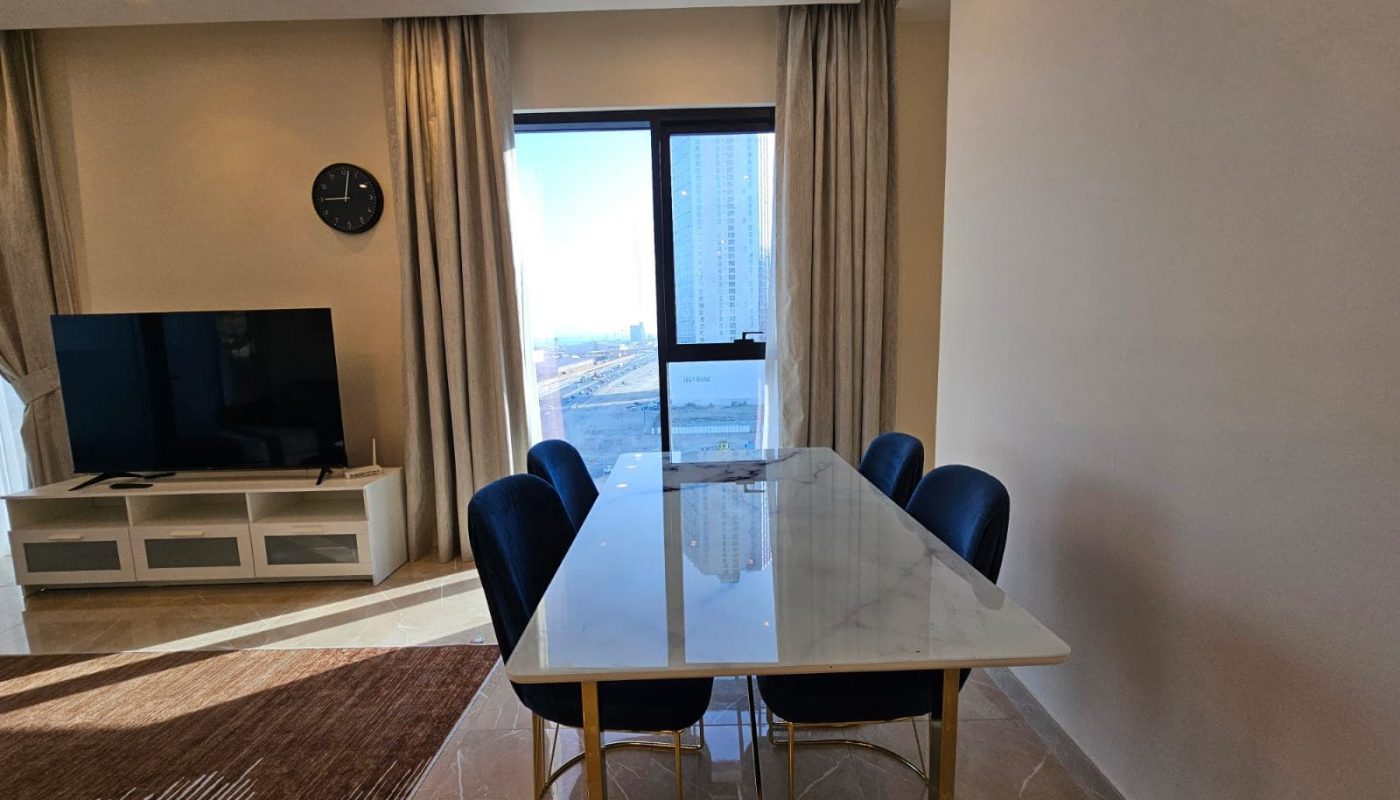 Interior of a modern living room featuring a marble dining table with blue chairs, a tv on a white stand, and a view of the city through a balcony door.