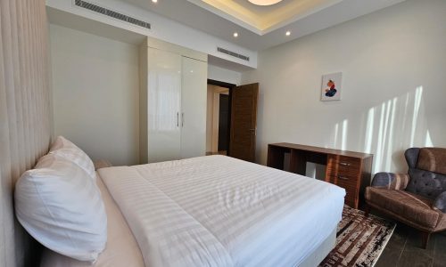 Modern bedroom with a large bed, white bedding, a wooden desk, brown armchair, and a small colorful artwork on the wall.