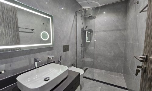 Modern bathroom interior with gray tiled walls, a glass shower, illuminated mirror, and a white vessel sink.