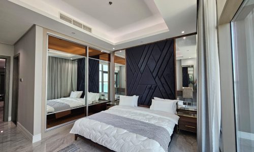 Modern hotel room with a large bed, reflective sliding closet doors, and an adjoining bathroom visible through a glass partition.