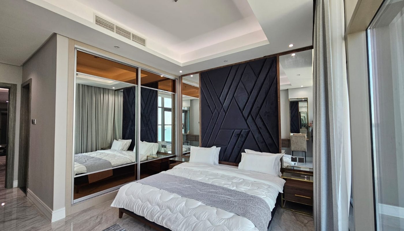 Modern hotel room with a large bed, reflective sliding closet doors, and an adjoining bathroom visible through a glass partition.