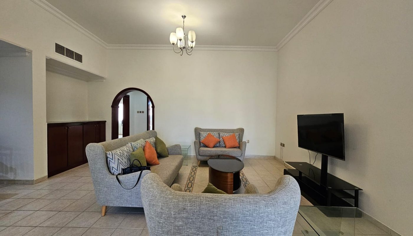 Spacious living room in Juffair area flat for rent, featuring two sofas, a glass coffee table, and a wall-mounted TV, with tiled flooring and a white ceiling light.