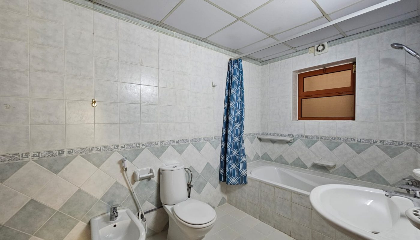 A small, tiled bathroom in a Juffair Area flat featuring a bathtub with a blue shower curtain, a toilet, bidet, sink, and a wooden cabinet above the sink.