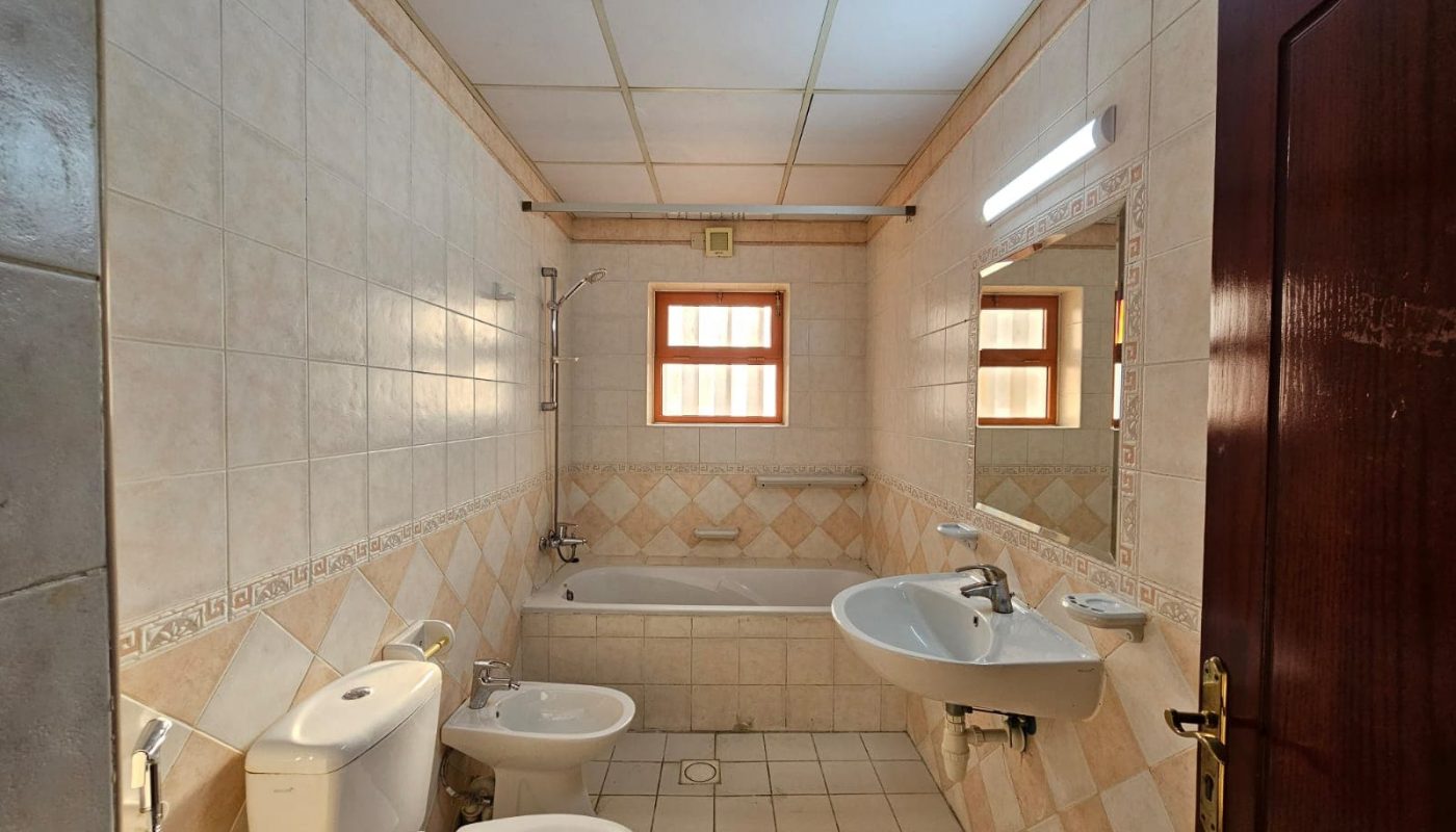 A clean bathroom in a flat for rent, featuring a toilet, bidet, sink, and bathtub, with tiled walls and floors, and two small windows that provide natural light.