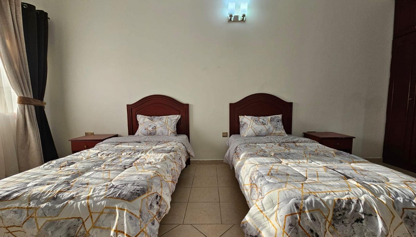A basic flat for rent with two single beds covered in patterned bedding, tile floor, and a small wall-mounted light.