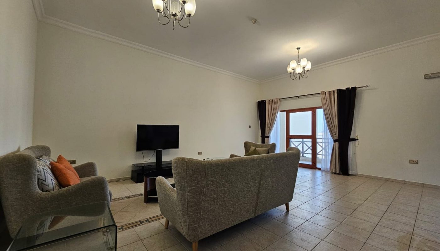Spacious living room with two gray sofas, a glass coffee table, tile flooring, and elegant light fixtures in Juffair Area, leading to a balcony with curtains.