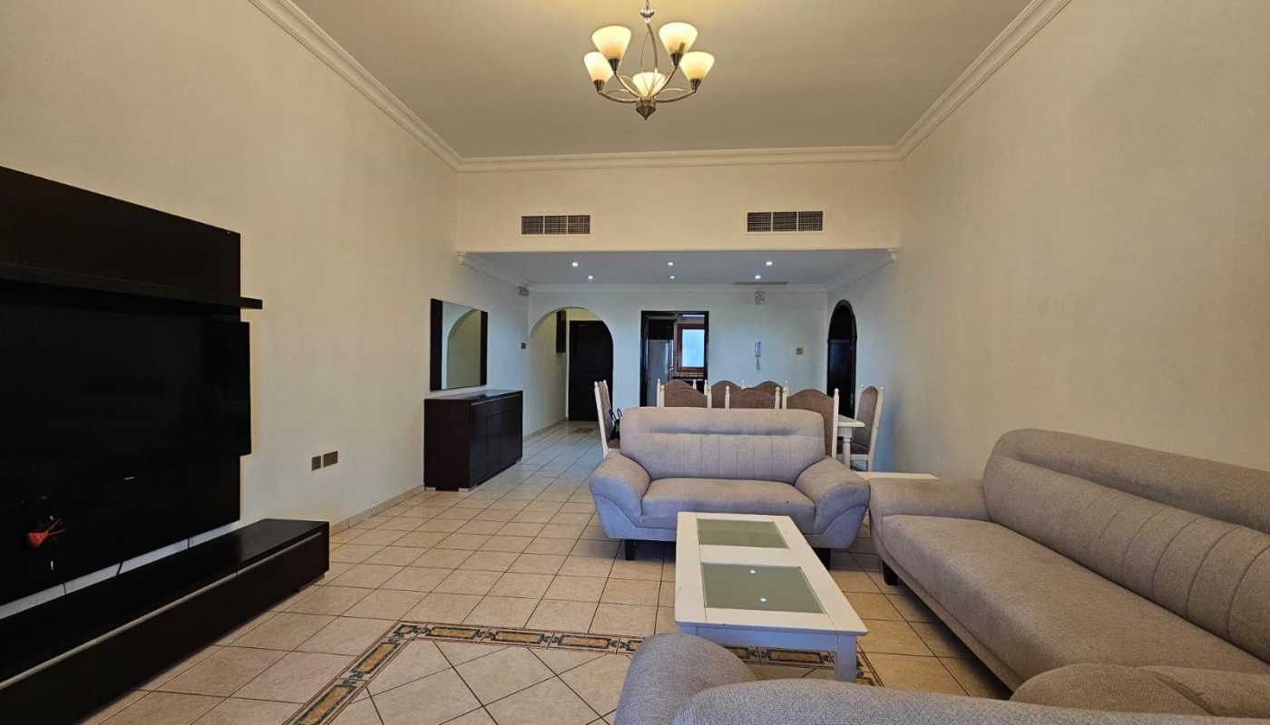 Spacious living room in a flat for rent in Juffair with beige walls, large gray couches, and a white coffee table, featuring a mounted flat-screen TV and arched doorways.