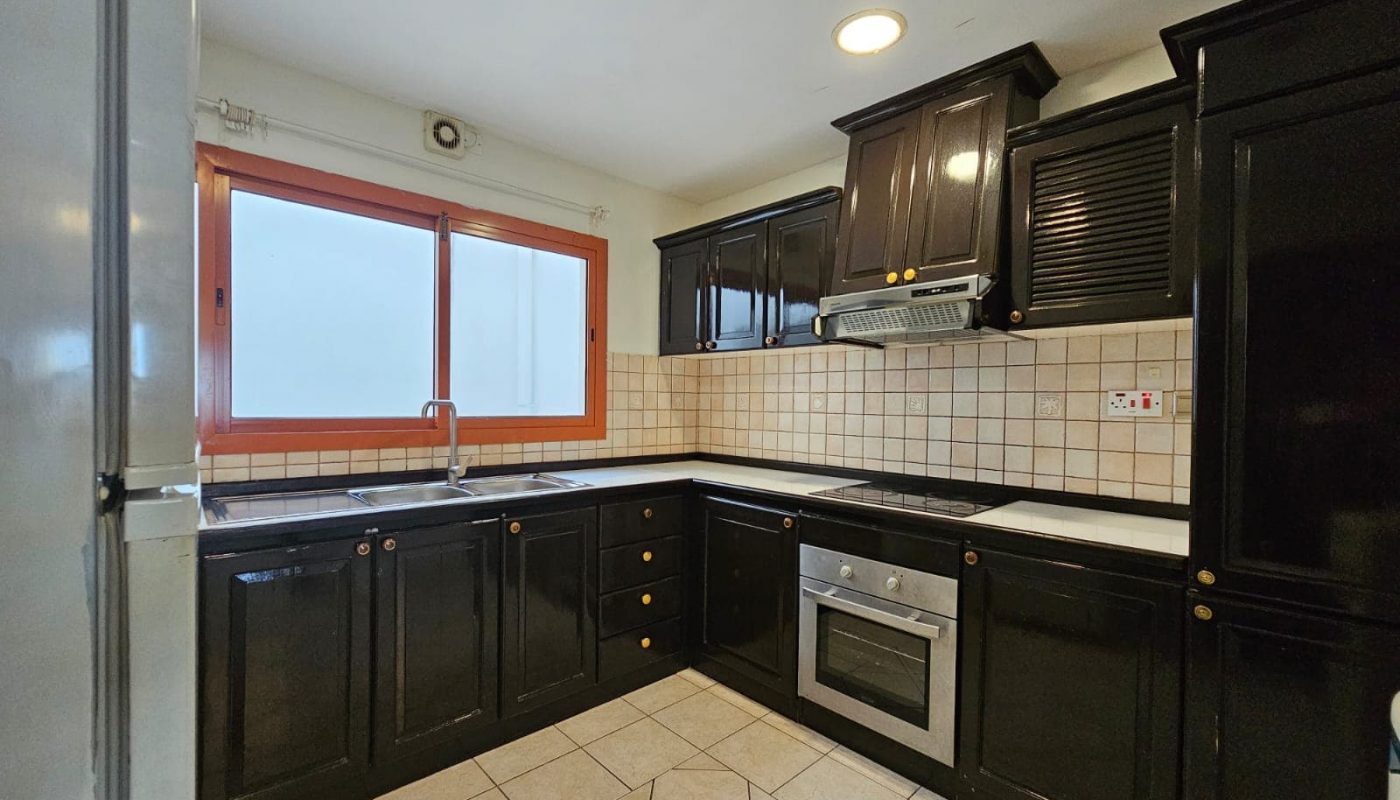 A kitchen in a Juffair flat for rent with black cabinets, white tiled backsplash, and built-in appliances, featuring a large window over the sink.