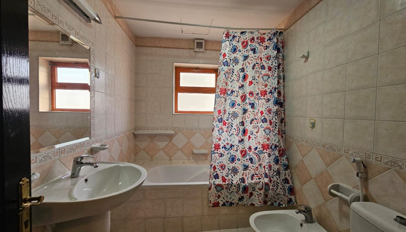 A small bathroom with beige tiles, in a flat for rent in Juffair, featuring a bathtub with a colorful curtain, a sink, toilet, bidet, and two small windows.