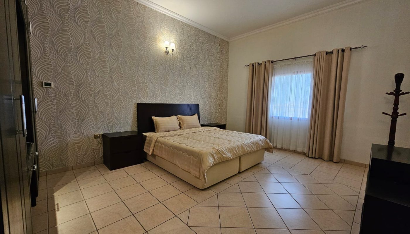 A modern bedroom with a double bed, beige bedding, patterned wallpaper, tiled floor, curtains, and minimalistic furniture is available in a flat for rent in Juffair.