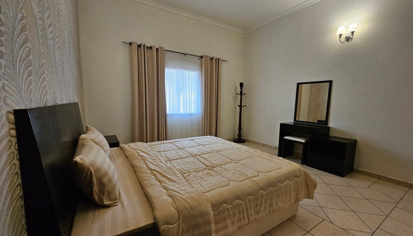 A simple bedroom in a Juffair flat for rent with a beige quilted bed, a wooden headboard, and minimalistic furnishings including a mirror and a coat stand.
