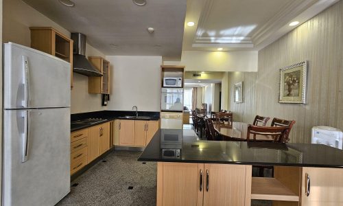 A modern kitchen and dining area in this Juffair flat for rent features a large refrigerator, wooden cabinetry, black countertops, a microwave oven, and a dining table with chairs. The space is well-lit and neatly arranged.