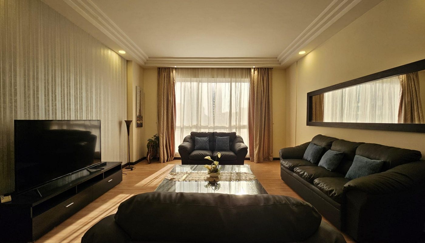 A modern living room in this Juffair rental property features dark couches, a large flat-screen TV, a glass coffee table, a large wall mirror, and ceiling lights. Sunlight streams through sheer curtains covering a large window. Perfect for anyone looking for the ideal flat for rent.