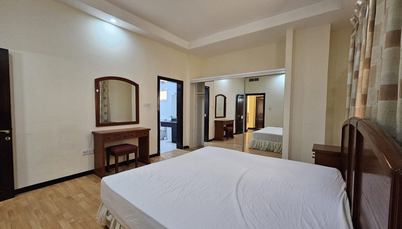 A sparsely furnished bedroom in a Juffair rental property, featuring a bed, mirrored vanity, chair, and light-colored walls. There are two doorways, one leading to what appears to be a bathroom. Ideal flat for rent seekers looking for simplicity and comfort.