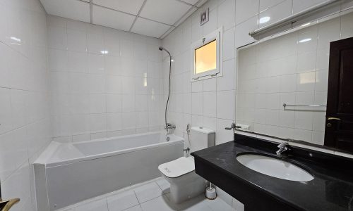 A pristine, white-tiled bathroom in a Juffair flat for rent features a bathtub with a handheld showerhead, a toilet, a sink with a black counter, and a mirror. The ceiling boasts rectangular tiles and there's a small window allowing natural light.