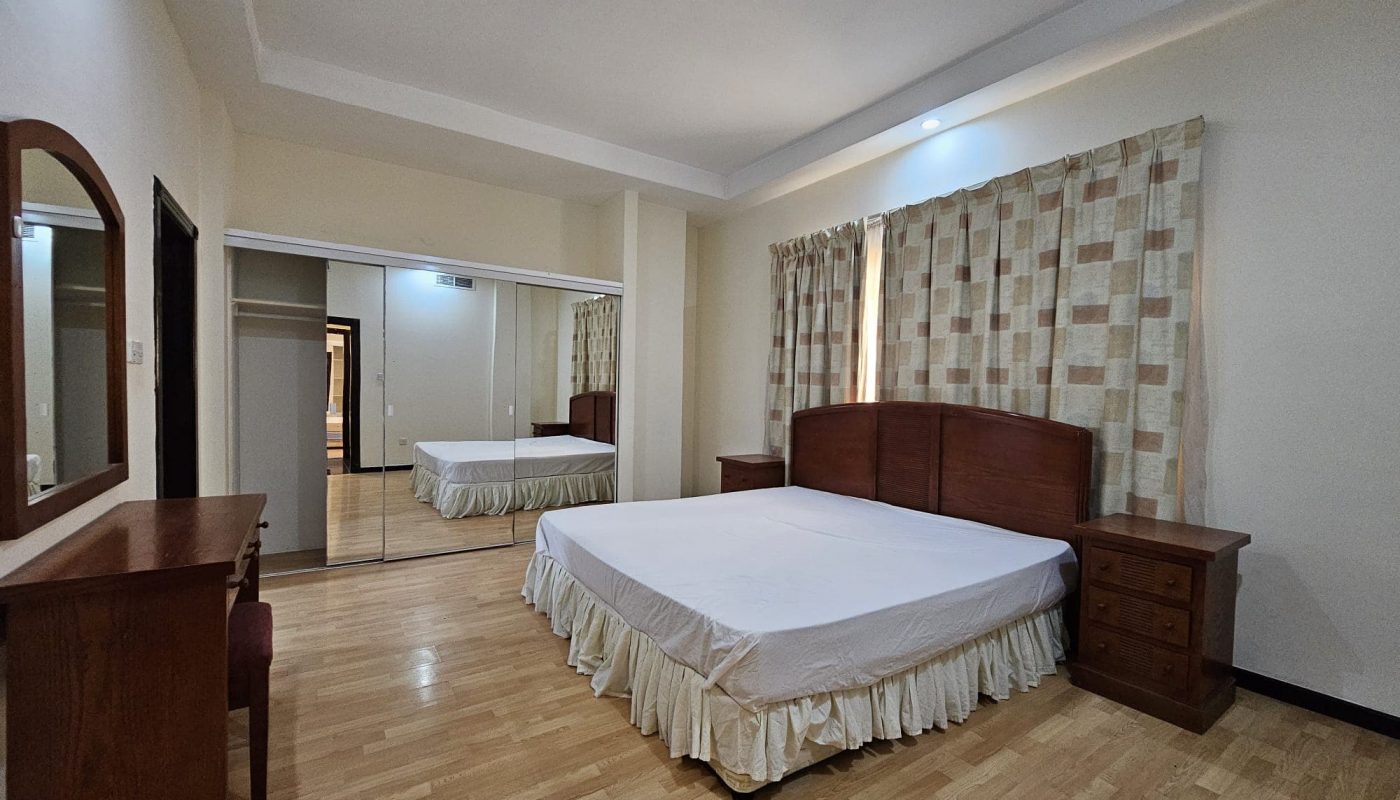 This rental apartment in Juffair features a bedroom with a double bed, wooden headboard, two bedside tables, a large mirror, and a wooden dresser with an attached mirror. The room boasts wooden flooring and patterned curtains, making it an ideal flat for rent.