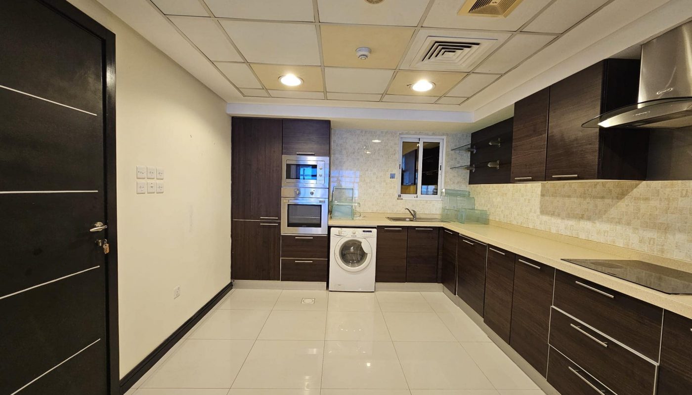 Modern kitchen with dark brown cabinets, white appliances including an oven, microwave, and washing machine, beige countertops, white tiled floors, and recessed lighting in a flat for rent in Juffair.