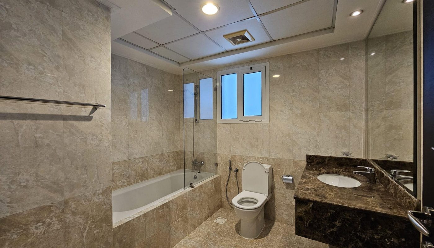 Modern bathroom in a Juffair flat for rent, featuring marble walls and flooring, a bathtub, toilet, and a sink with a marble countertop. A large mirror and windows provide natural light.