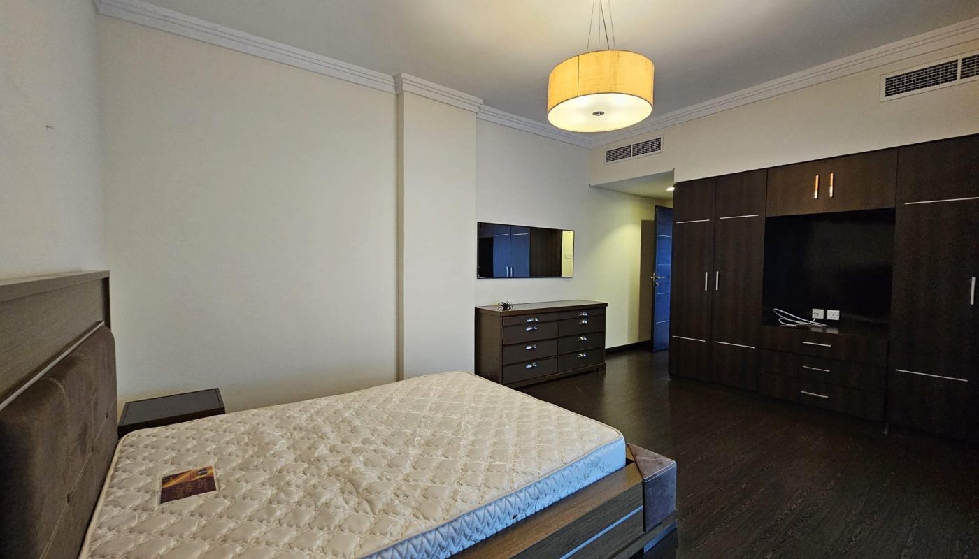 A flat for rent in Juffair features a bedroom with a single bed, wooden furniture, and a ceiling light fixture. The room includes a wardrobe, dresser, and a wall-mounted television. The bed has a mattress without bedding.
