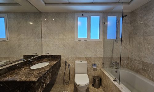 A flat for rent in Juffair offers a bathroom featuring a marble countertop with a sink, a toilet, a glass-enclosed bathtub, and frosted windows.