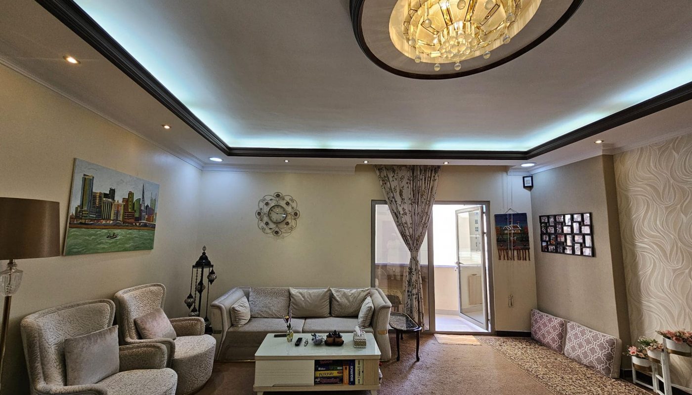 A well-lit living room in this modern flat in Juffair features a sleek chandelier, a wall-mounted painting, a grey sofa, two armchairs, a central coffee table, and a sliding glass door leading to an outdoor space. Available for rent now.