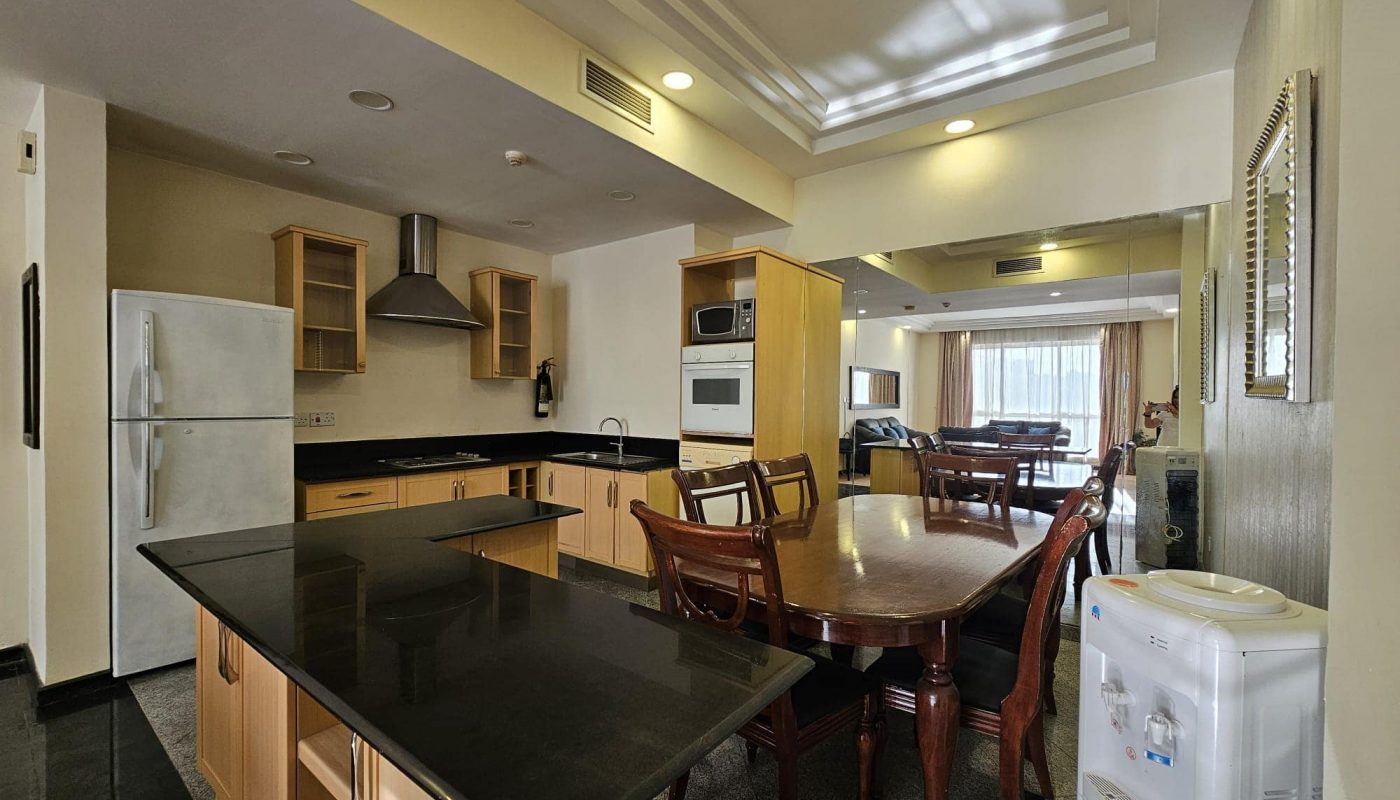 Modern kitchen and dining area with a central island, granite countertops, stainless steel appliances, wooden cabinets, and a water dispenser. The background shows a living room with a sofa and large windows. This stylish flat for rent in Juffair is perfect for anyone looking to rent property in the area.