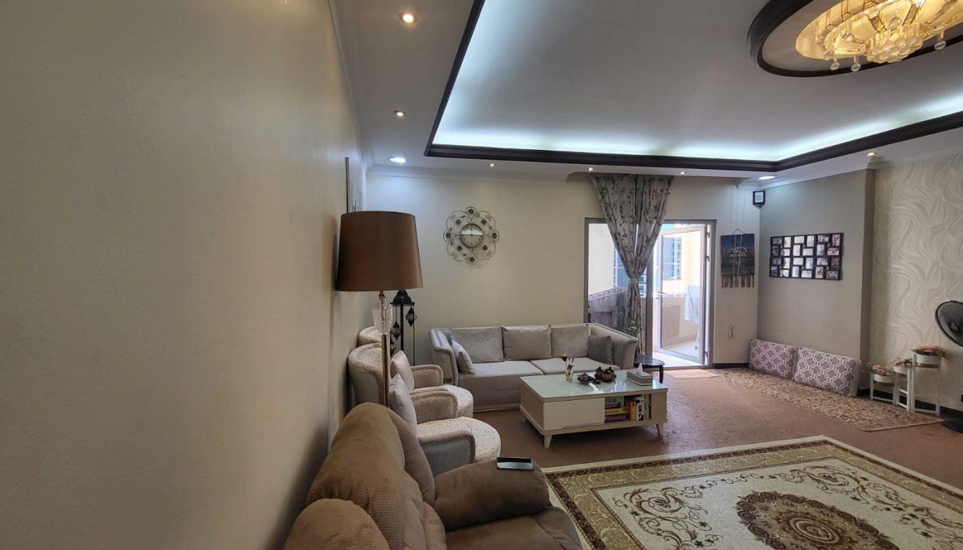 A spacious living room in a modern flat with multiple sofas, a coffee table, a floor lamp, a wall clock, and a large rug. The room leads to a balcony through a glass door with curtains. A chandelier adorns the ceiling. Located in Juffair and available for rent.