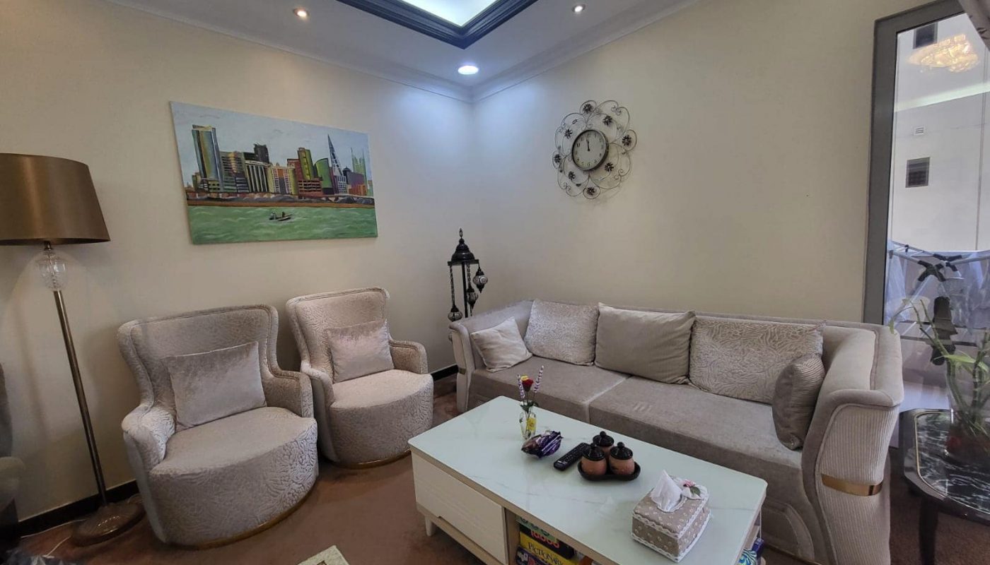 A cozy living room in a modern flat for rent in Juffair, featuring a white sofa, two beige swivel chairs, a coffee table with books, a wall clock, and a cityscape painting. A tall floor lamp and a side lantern add decorative elements.