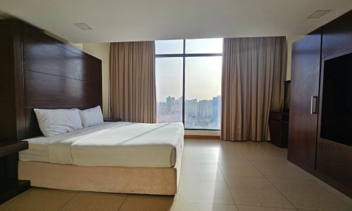 A flat for rent in Juffair offers a hotel room with a large bed, light brown tile floor, wooden furnishings, tall windows with drawn beige curtains, and a cityscape view in the background.