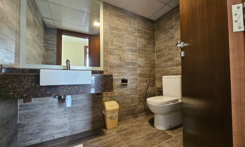 A modern bathroom in a flat for rent features a sink and wall-mounted mirror on the left, a toilet on the right, and a small trash bin placed under the sink. The walls and floor are tiled in brown, making it an elegant space to rent in Juffair.