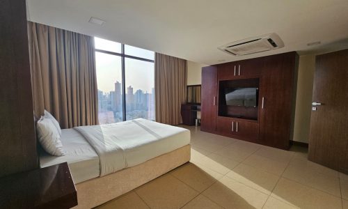 A modern hotel room in Juffair with a large bed, wooden furniture, floor-to-ceiling windows, and a view of the city skyline. The room features beige tiles and beige curtains, perfect for those seeking a flat for rent in Juffair.