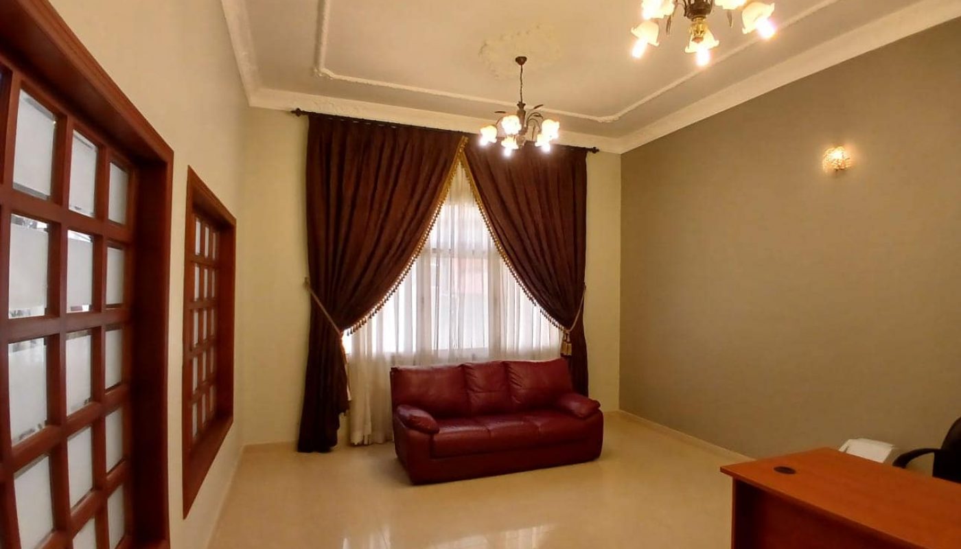 A commercial villa features a living room with beige tiled floors, brown curtains, a red couch, a wooden desk, chair, and two ceiling light fixtures available for rent.