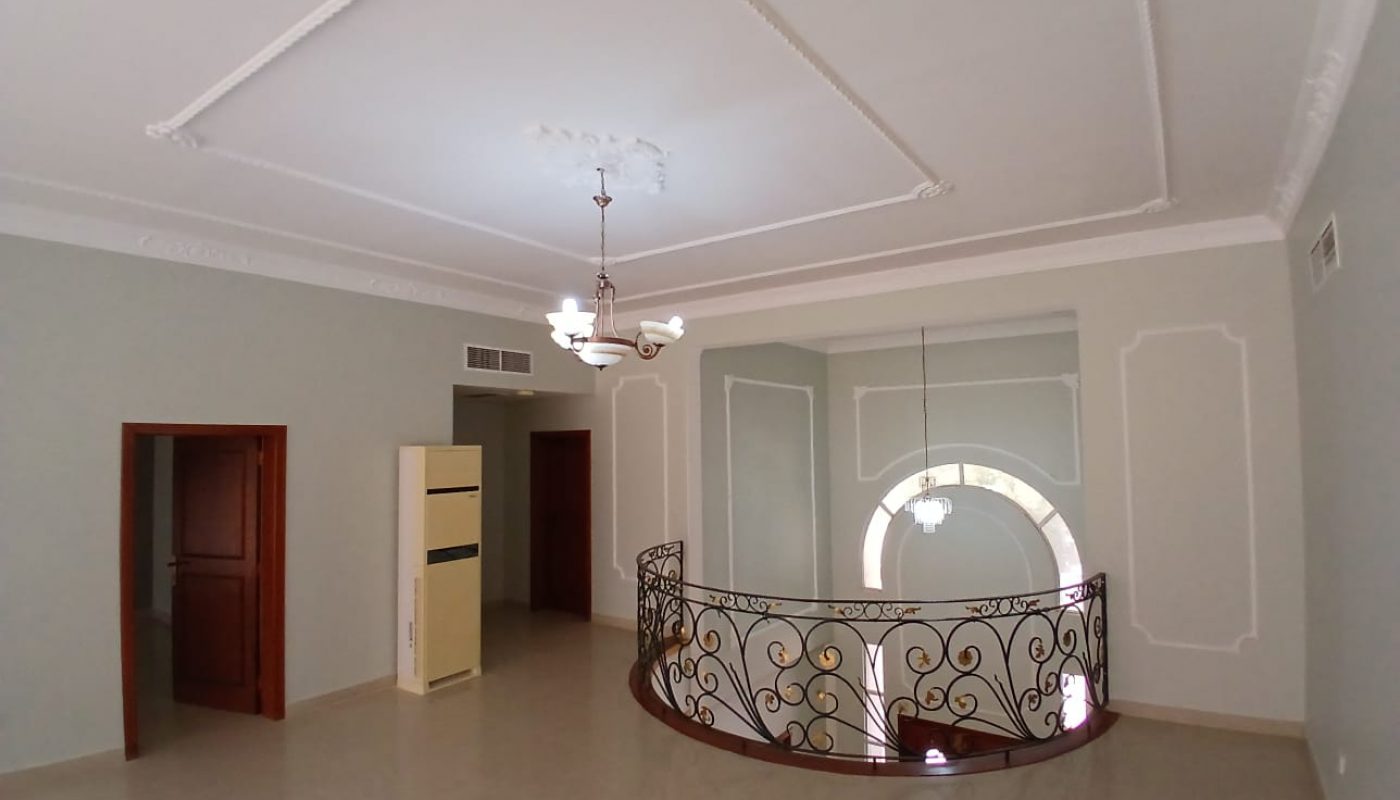 A spacious, unfurnished room in this commercial villa features a chandelier, ornate ceiling moldings, and a spiral staircase leading to a lower level. The room has beige walls and tile flooring, ideal for rent as an elegant office or boutique space.