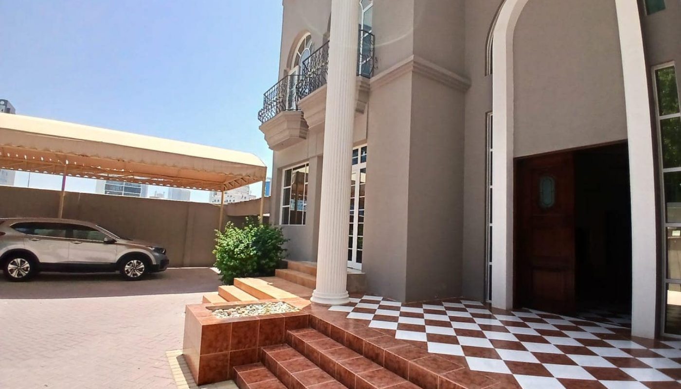 A modern commercial villa with gray walls and white columns. The entrance features a brown tiled staircase, ideal for professional settings. A car is parked under a beige canopy to the left, making it convenient for tenants and visitors alike. Perfect for rent!