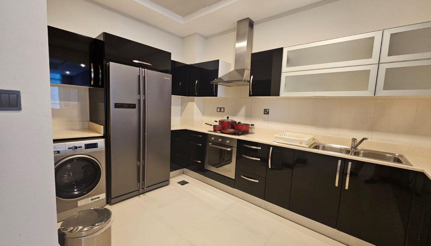 Modern kitchen with black cabinets, stainless steel fridge, oven, washing machine, and hood. Includes a double sink with a faucet and a dish rack. White tiled floors and walls with overhead lighting.