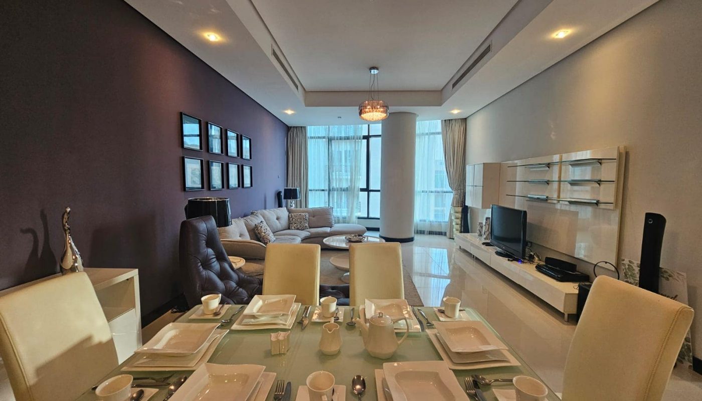 A modern, well-lit living and dining area with a glass dining table set for six, contemporary decor, a chandelier, a wall-mounted TV, and large windows covered with sheer curtains.