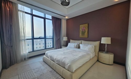 A modern bedroom with a large bed, white linens, two bedside tables with lamps, and a window with sheer curtains, showcasing a view of buildings outside. The walls are painted in neutral tones.