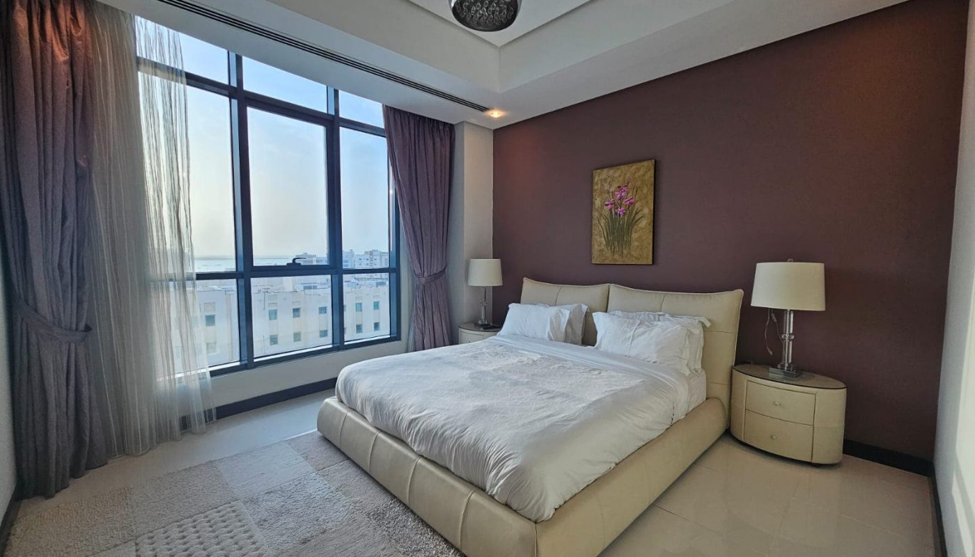 A modern bedroom with a large bed, white linens, two bedside tables with lamps, and a window with sheer curtains, showcasing a view of buildings outside. The walls are painted in neutral tones.