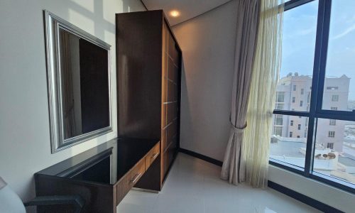 A sunlit corner of a modern room with a large window, long beige curtains, a wall-mounted mirror, a dark wooden cabinet, and a glass-topped desk.