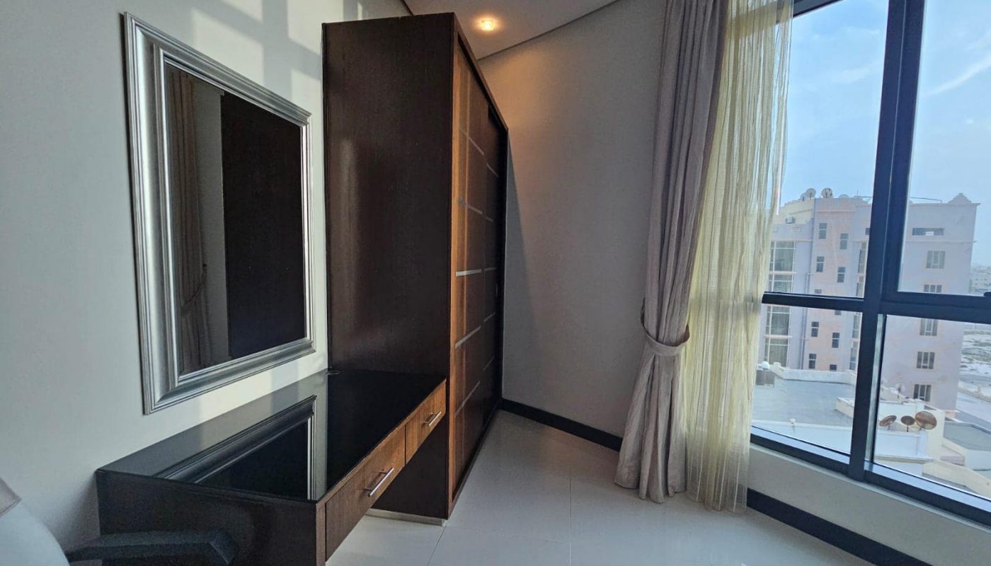 A sunlit corner of a modern room with a large window, long beige curtains, a wall-mounted mirror, a dark wooden cabinet, and a glass-topped desk.