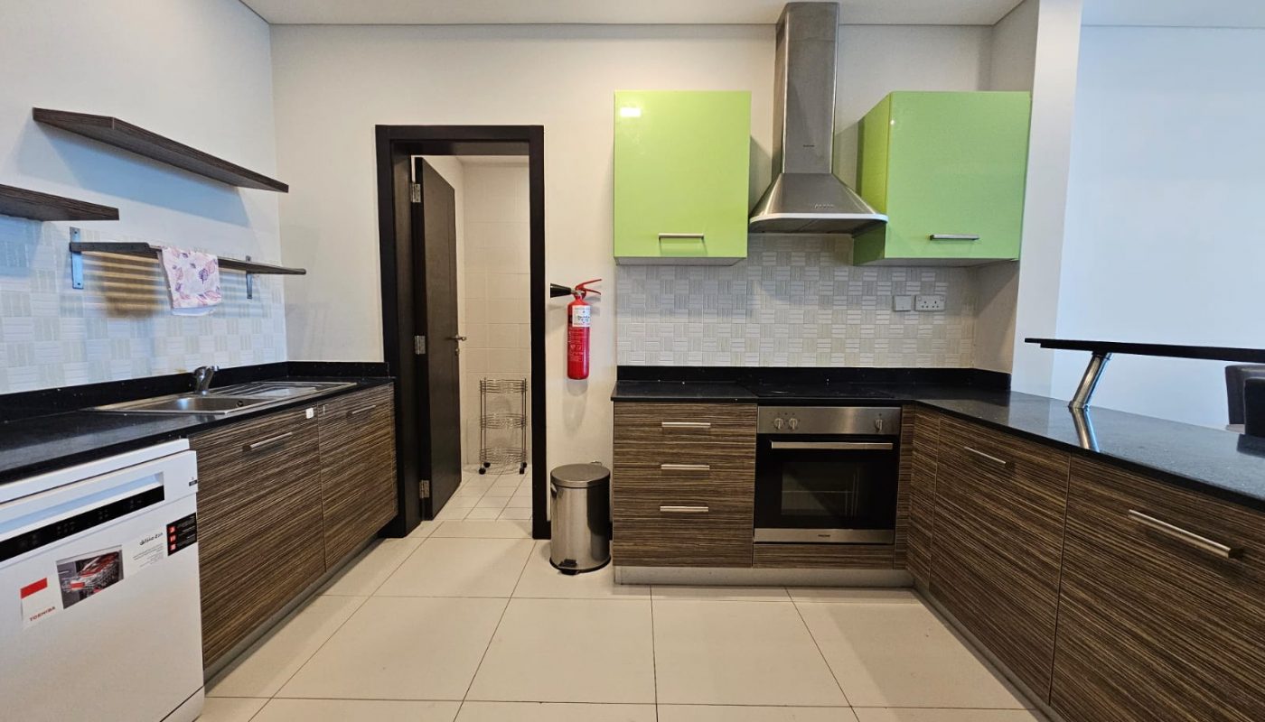 Modern kitchen with green upper cabinets, wooden lower cabinets, stainless steel appliances, black countertops, a fire extinguisher on a white tile wall, and an open doorway leading to another room. This stylish setup is part of a chic flat for rent on Reef Island.